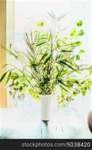 Lovely plants bunch in vase on table at window background. Florist arrangements with variety of green tropical plants.Home decor and interior, backlight