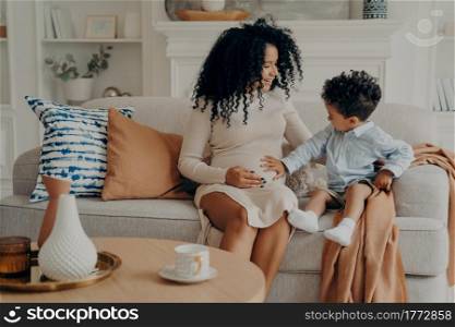 Lovely pregnant mixed race woman mother explaining to curious son about his future brother or sister, cute little boy touch belly of his mom and talking with baby while sitting on couch in living room. Cute little boy touching belly of his pregnant mom while relaxing together on sofa in living room
