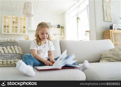 Lovely preschool child is learning to read. Little european girl is sitting on sofa and reading book. Kid is studying at home on quarantine. Leisure and study at cozy home on quarantine.. Little european girl with book has leisure at cozy home. Preschool child is learning to read.