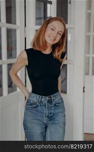 Lovely redhead caucasian model in bodysuit and jeans is standing near door window and smiling. Portrait of positive blonde young woman. Concept of glamour, style, fashion and beauty.. Lovely redhead caucasian model in bodysuit and jeans is standing near door window and smiling.