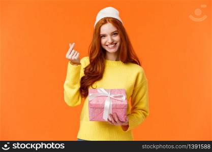 Lovely redhead girlfriend got gift from her girl, showing korean heart gesture and smiling silly, holding pink present box, wearing winter beanie, standing over orange background happy.. Lovely redhead girlfriend got gift from her girl, showing korean heart gesture and smiling silly, holding pink present box, wearing winter beanie, standing over orange background happy
