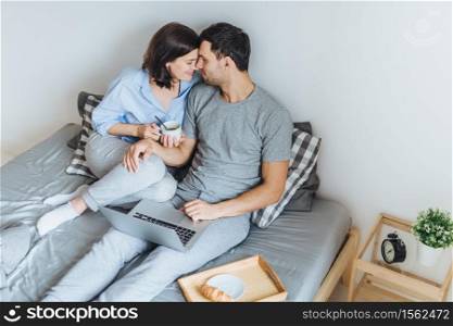 Lovely romantic couple going to kiss, look at each other as sit on bed, use laptop computer for watching films, drink coffee, enjoy togetherness and relaxation in bedroom. Relationship concept