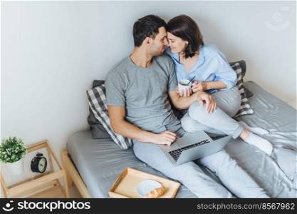 Lovely romantic couple going to kiss, look at each other as sit on bed, use laptop computer for watching films, drink coffee, enjoy togetherness and relaxation in bedroom. Relationship concept