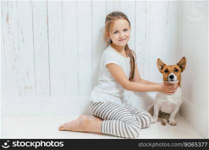 Lovely small female child plays with her dog in white room, sit on floor, have good relationship, cuddles favourite pet. Little schoolgirl likes animals, spends leisure time at home. Childhood