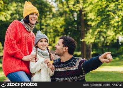 Lovely small girl with two pigtails, wears knitted hat, scarf and sweater, holds leaf in hand, looks joyfully into distance as her father shows her beautiful flower. Spending autumn days together