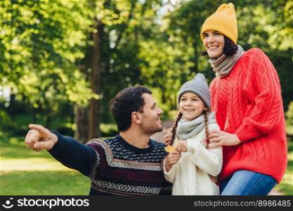 Lovely small girl with two pigtails, wears knitted hat, scarf and sweater, holds leaf in hand, looks joyfully into distance as her father shows her beautiful flower. Spending autumn days together