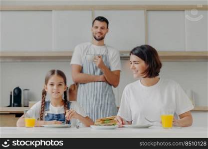 Lovely small kid and mother have breakfast together, sit at kitchen table, eat delicious meal, father stands in background, wears apron and drink coffee. Friendly family gather together at kitchen