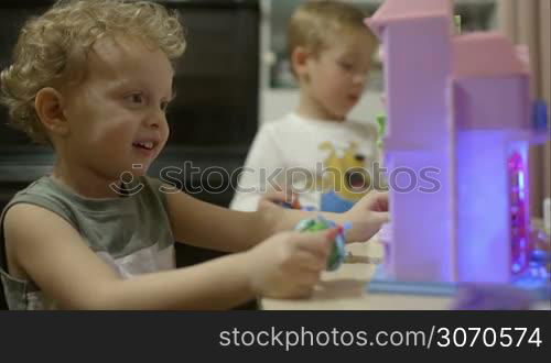 Lovely smiling little boy playing with toy plastic house, another child playing near him. Happy childhood