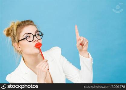 Lovely sweet business woman elegant clothing nerdy glasses holding red fake lips on stick having fun, pointing up with finger at copy space, on blue. Carnival funny accessories concept.. woman holds fake lips on stick pointing at copy space