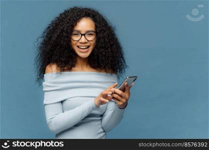 Lovely teenage girl enjoys online communication, holds modern cell phone, checks email box, connected to wireless internet, cannot imagine life without technologies, isolated on blue studio wall