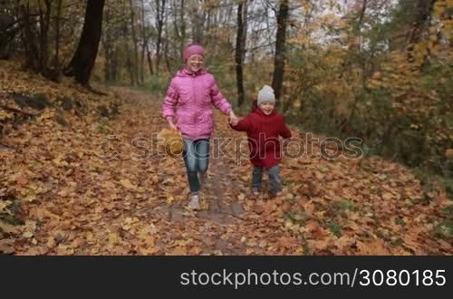 Lovely teenage girl in eyeglasses carrying yellow maple leaves and her cute toddler brother running through autumn park. Joyful siblings enjoying freetime and having fun, running on park road over golden autumn landscape background. Slo mo.