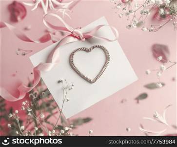 Lovely white greeting card with heart at pink background with petals, ribbon and leaves.