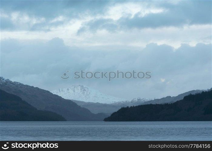 Lovely Winter landscape image of view along Loch Lomond towards snowcapped mountain range in distance during blue hour