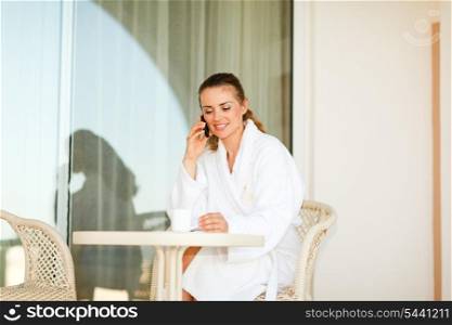 Lovely woman in bathrobe sitting at table on terrace having coffee and speaking phone&#xA;