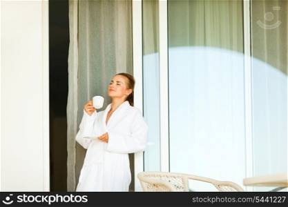 Lovely woman in bathrobe standing near big room windows and having cup of coffee