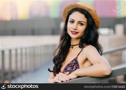 Lovely woman with dark long hair, appealing appearance wearing summer hat and dress having excursion in big city posing at bridge. Young female traveler walking on street having delightful look