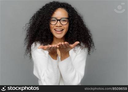 Lovely young African American woman sends air kiss, keeps palms stretched near mouth, shows white teeth, expresses love to somebody, wears spectacles and sweater, isolated over grey background.