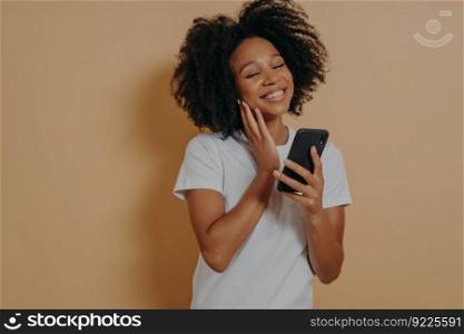 Lovely young african american woman with modern smartphone in hands feeling happy to get message from her boyfriend, looking at device screen and smiling while standing over beige studio wall. Young african american woman with smartphone in hands feeling happy to get message from boyfriend