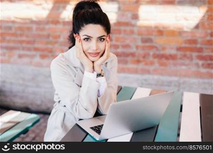 Lovely young brunette woman with charming face and red manicure wearing white coat sitting at table using laptop computer chatting with her family using free internet connection with high speed