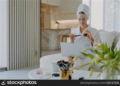 Lovely young Caucasian woman wears bathrobe and towel on head holds cosmetic brush in front of laptop computer gives tutorail lesson how to do makeup sits at comfortable sofa in modern apartment