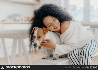 Lovely young curly African American woman embraces beloved pedigree dog with love, has gentle smile, wears stylish clothing, poses against home background in modern apartment, expresses care