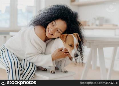 Lovely young curly African American woman embraces beloved pedigree dog with love, has gentle smile, wears stylish clothing, poses against home background in modern apartment, expresses care