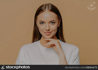 Lovely young European woman with makeup healthy skin touches gently jawline looks directly at camera has long straigh hair natural beauty dressed in casual sweater isolated over brown background