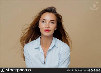 Lovely young female has long wavy hair floating in wind, dressed in elegant blue shirt, wears lipstick and makeup, poses against brown background, has direct gaze at camera, healthy facial skin