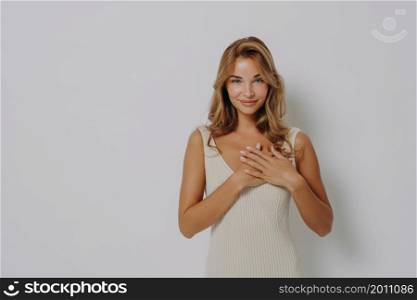 Lovely young female model presses palms to heart expresses gratitude says thank you wears dress applies minimal makeup poses against grey background with copy space for your promotional text. Lovely young female model presses palms to heart expresses gratitude says thank you