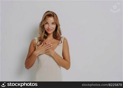 Lovely young female model presses palms to heart expresses gratitude says thank you wears dress applies minimal makeup poses against grey background with copy space for your promotional text. Lovely young female model presses palms to heart expresses gratitude says thank you