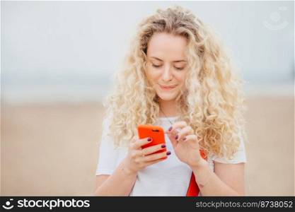 Lovely young female with curly bushy hair, healthy clean skin, holds modern smart phone, messages with friends in social networks, connected to wireless internet, strolls across seaside outdoor