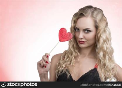 lovely young girl with wavy blonde hair-style and sexy dress posing with red heart shaped lollipop in the hand