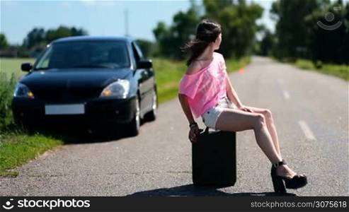 Lovely young woman on high heels is sitting on old canister in the centre of country road and looking for passing by traffic to ask assistance, on the background blurred car parked on roadside.