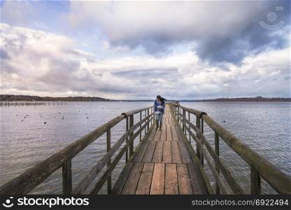 Lovely young woman walking alone on a long wooden bridge, over the famous Chiemsee lake, enjoying the breeze and the cold sunset of November, in Germany.