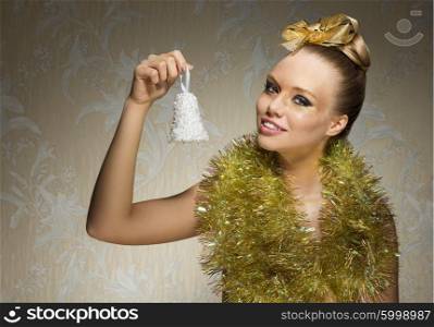 lovely young woman with freckles and creative shiny make-up posing in christmas artistic shoot with golden ribbon in hair-style, sparkly tinsel around neck and little bell in the hand