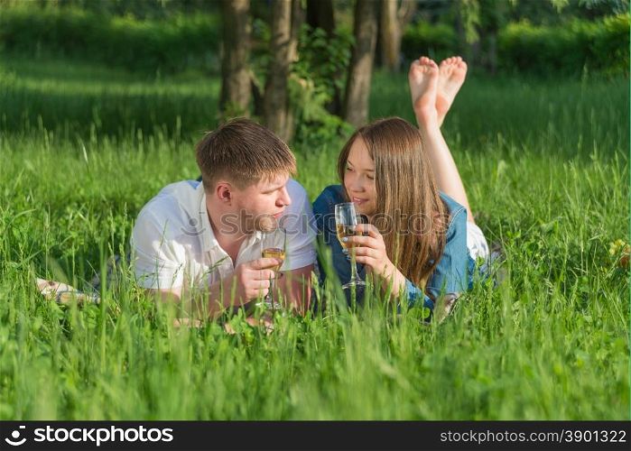 Lovers girl and boy lying on the grass and drinking white wine during a picnic in a city park