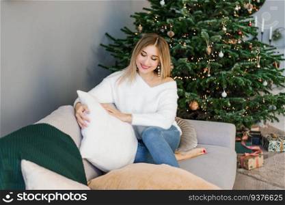 Lovey woman is sitting on a gray sofa near the Christmas tree in the living room and holding a soft pillow in her hands and smiling