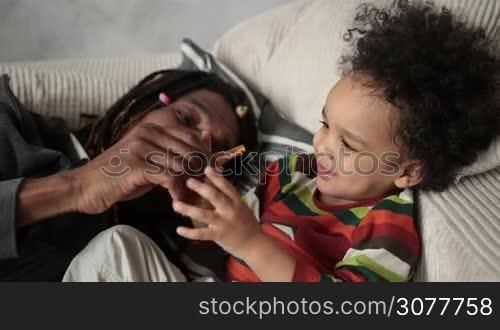 Loving african american dad and mixed race toddler son playing and having fun while lying on the sofa at home closeup. Cheerful father fooling around trying to pinch nose of laughing child with clothespin. Slow motion. Top view.