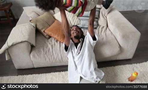 Loving african american dad with dreadlocks lifting his curly mixed race son up into the air. Stylish hipster father sitting on the floor and raising his cute giggling child up high in modern apartment. Slow motion.