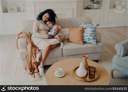 Loving african pregnant mom tenderly embracing little son while sitting on cozy couch next to coffee table in living room, spending leisure time together at home during weekend. Motherhood concept. Single african pregnant mom tenderly embracing little son while sitting on cozy couch