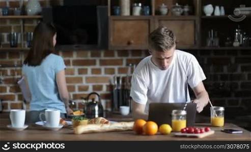 Loving couple enjoying great time together while having breakfast in modern kitchen in the morning. Handsome man chatting about latest news with her cute girlfriend. Beatiful brunette girl pouring some tea into cup and offering to her boyfriend.