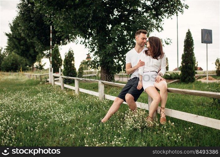 Loving couple is sitting in the summer park on white fence. man and woman are hugging outdoors on warm day.. Loving couple is sitting in the summer park on white fence. man and woman are hugging outdoors on warm day