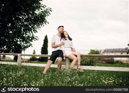Loving couple is sitting in the summer park on white fence. man and woman are hugging outdoors on warm day.. Loving couple is sitting in the summer park on white fence. man and woman are hugging outdoors on warm day