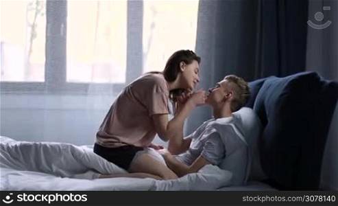 Loving couple lying on the bed, hugging each other and gently kissing after waking up in bright modern bedroom at home. Passionate beautiful couple in bedroom enjoying leisure in the morning. Side view.