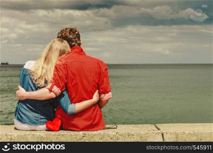 Loving couple spending leisure time together at beach sitting on sea shore with red heart hugging rear view