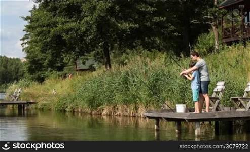 Loving father teaching his teenage boy how to fish with spinning rod and real on the lake over amazing rural landscape background. Positive dad helping son to cast fishing rod while fishing together at freshwater pond on sunny summer day.