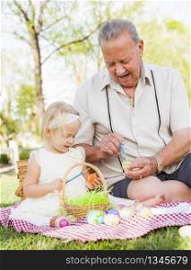 Loving Grandfather and Granddaughter Coloring Easter Eggs Together on Picnic Blanket At The Park.