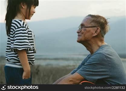 Loving grandfather and little granddaughter look at each other and smile against the sunset background. Family love and spending time outdoors together.