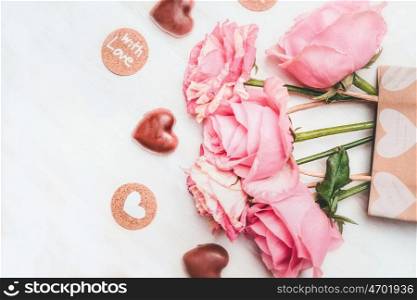 Loving greeting card with pink roses, chocolate with heart symbol and text with love on white wooden background, top view