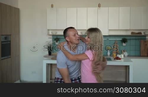 Loving handsome man carrying attractive wife in his arms and spinning around in the morning over modern styled domestic interior. Romantic husband in pajamas whirling his beloved woman in kitchen at home. Slow motion.